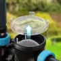 Nærbillede af gennemsigtigt plastfilterlåg, der løftes af sort cylindrisk filterhus med perforeret indvendig kurv. Bruges til vandfiltreringssystemer. Blå beslag ses på siderne.