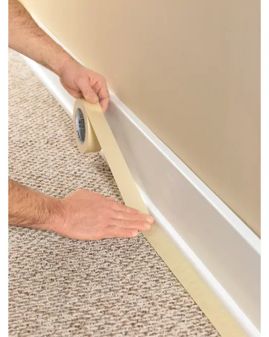 Beige malertape påføres langs kanten af et tæppe og fodpanel, perfekt til beskyttelse under maling eller renovering. Bred rulle for nem dækning.