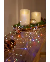 A string of decorative lights shaped like starbursts, each with multiple thin silver arms ending in small, colorful LED bulbs. The lights emit red, green, blue, and yellow hues, creating a festive atmosphere. The lights are displayed on a wooden surface alongside shiny Christmas ornaments and artificial candles, suggesting a holiday or celebratory setting.