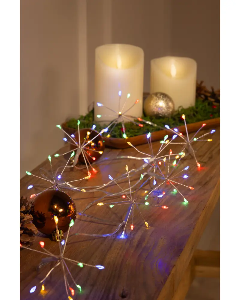 A string of decorative lights shaped like starbursts, each with multiple thin silver arms ending in small, colorful LED bulbs. The lights emit red, green, blue, and yellow hues, creating a festive atmosphere. The lights are displayed on a wooden surface alongside shiny Christmas ornaments and artificial candles, suggesting a holiday or celebratory setting.