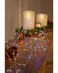 A string of decorative lights shaped like starbursts, each with multiple thin silver arms ending in small, colorful LED bulbs. The lights emit red, green, blue, and yellow hues, creating a festive atmosphere. The lights are displayed on a wooden surface alongside shiny Christmas ornaments and artificial candles, suggesting a holiday or celebratory setting.