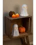 The image shows a rustic wooden crate used as a shelf, decorated for Halloween. On the top shelf, there is a glossy white ceramic ghost with cut-out eyes and mouth, a brown textured pumpkin, and two small orange pumpkin-shaped candles with lit flames. On the lower shelf, there is another smaller ceramic ghost with cut-out eyes and mouth, and a large orange pumpkin with a twisted stem. The overall arrangement creates a festive and cozy autumn atmosphere.