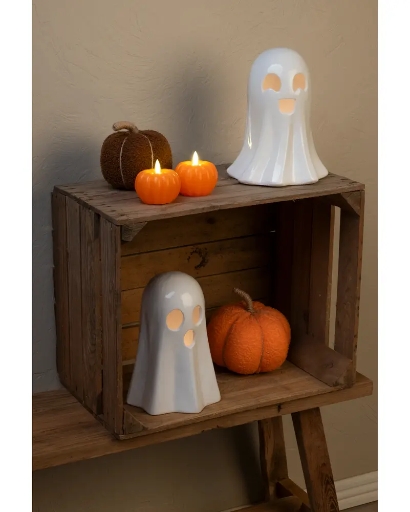 The image shows a rustic wooden crate used as a shelf, decorated for Halloween. On the top shelf, there is a glossy white ceramic ghost with cut-out eyes and mouth, a brown textured pumpkin, and two small orange pumpkin-shaped candles with lit flames. On the lower shelf, there is another smaller ceramic ghost with cut-out eyes and mouth, and a large orange pumpkin with a twisted stem. The overall arrangement creates a festive and cozy autumn atmosphere.