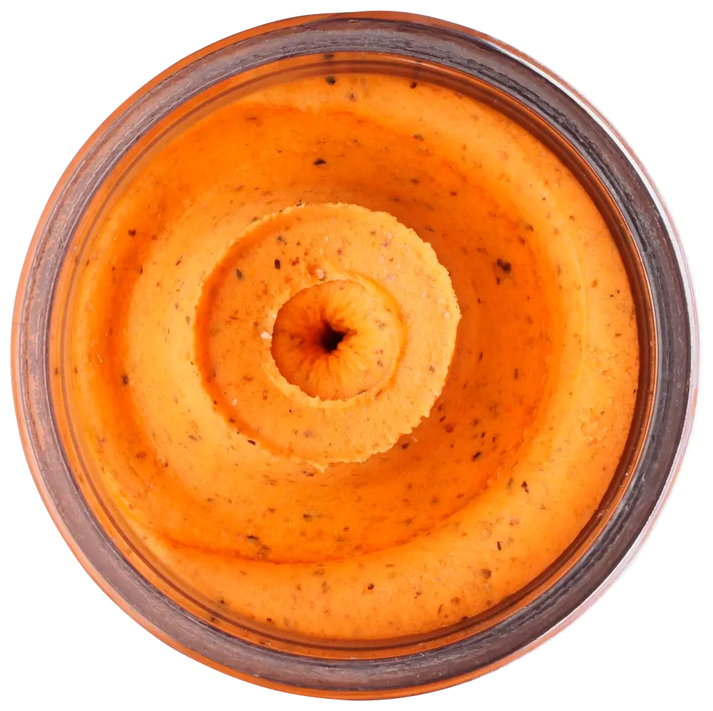 Slät, orangefärgad röra eller dipp i glasburk, med synliga örter och kryddor, krämig konsistens, troligen smaksatt hummus eller grönsaksbaserad röra.