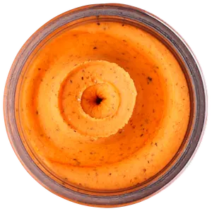 Slät, orangefärgad röra eller dipp i glasburk, med synliga örter och kryddor, krämig konsistens, troligen smaksatt hummus eller grönsaksbaserad röra.