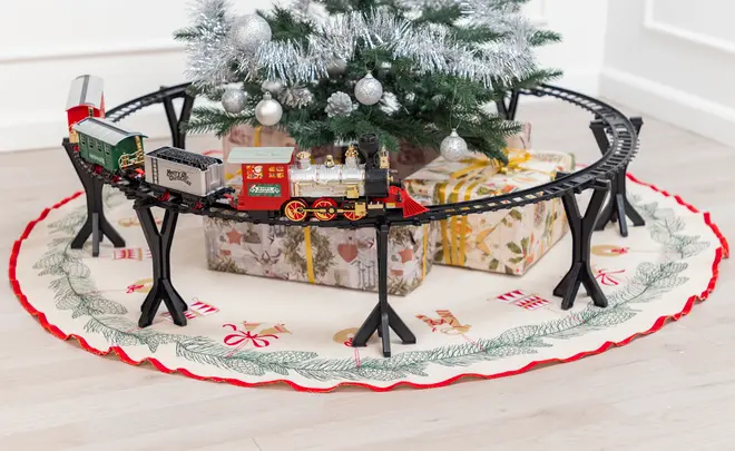 Elektriskt leksakståg med rött och guld lok, svart upphöjd räls och julgransmatta, placerat under en dekorerad gran med inslagna paket.