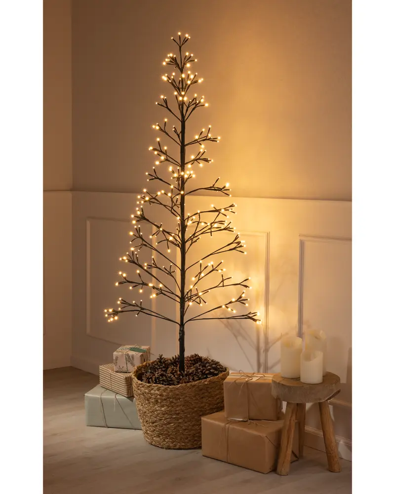 A minimalist artificial tree with bare branches, each adorned with small warm white LED lights. The tree is set in a woven basket filled with pinecones, giving it a natural look. It stands in a softly lit room, surrounded by wrapped gifts in neutral tones and a small wooden stool holding three pillar candles.