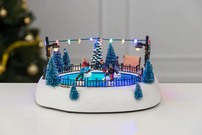 Miniatyrskridskobana med två åkare, snötäckta granar, bänk, svart staket och färgglada ljusslingor på vit snöbas. Dekorativ julprydnad.