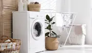 Frontmatad vit tvättmaskin med genomskinlig lucka och kontrollpanel ovanpå, placerad i ljust tvättrum. Omgiven av tvättkorgar, torkställning och dekorativ växt.