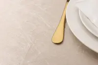 Elegant smörkniv i guldfärg bredvid vit keramiktallrik med vikt vit servett, placerad på ljusbeige texturerad bordsduk.