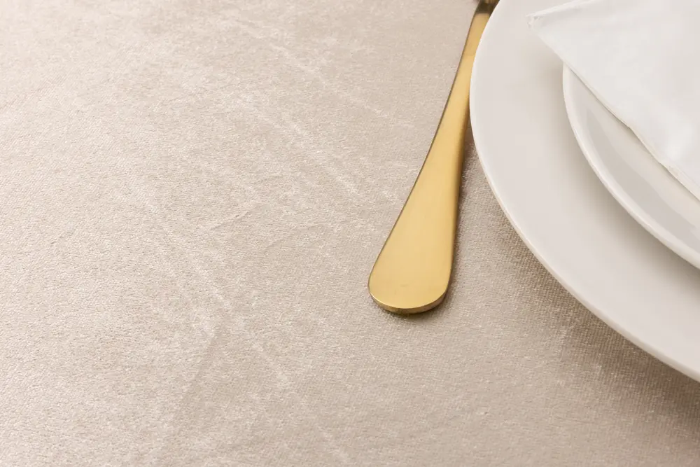 Elegant smörkniv i guldfärg bredvid vit keramiktallrik med vikt vit servett, placerad på ljusbeige texturerad bordsduk.