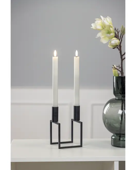 Modern svart metall-ljusstake med geometrisk fyrkantig bas, rymmer två höga vita kronljus. Minimalistisk design, ljusen är tända på bilden.