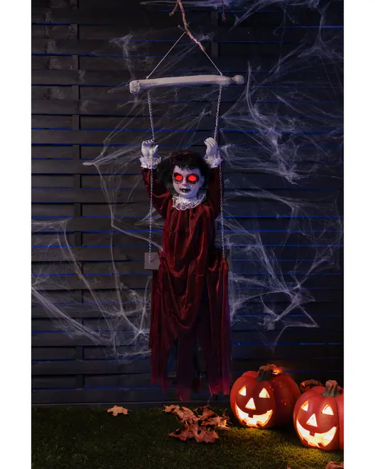 Hängande Halloween-dekoration med skrämmande marionettdocka, lysande röda ögon, röd klänning, omgiven av konstgjorda spindelnät och två upplysta pumpalyktor.