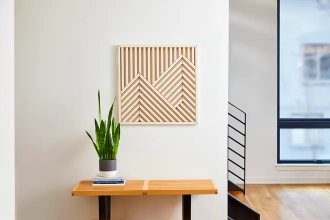 Modern väggkonst i trä med geometriskt bergmotiv i ljusa och mörka trätoner. Hänger ovanför ett ribbat bord med krukväxt och staplade böcker.