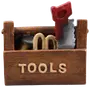 Brun værktøjskasse med "TOOLS" på fronten, indeholder små værktøjer som en sav med rød håndtag og andre genstande. Fremstillet af malet materiale, dekorativ eller sjov brug.