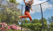 Udendørs trampolin med sikkerhedsnet, sort ramme og netvægge. Velegnet til børn, giver sikker hoppe- og legeplads. Vist i have med blomster og grønne planter.