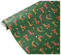 Green wrapping paper featuring a repeating pattern of dachshund dogs wearing red and white Christmas accessories such as Santa hats, scarves, and sweaters. Some dogs are holding gifts or have reindeer antlers. The design includes small stars, snowflakes, and the phrase 'Happy Xmas' scattered throughout.