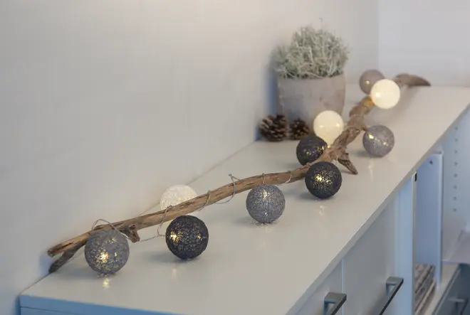 Dekorativ ljusslinga med svarta, grå och vita klot, fästa på en rustik trägren, perfekt för heminredning eller fest, placerad på ett vitt skåp.