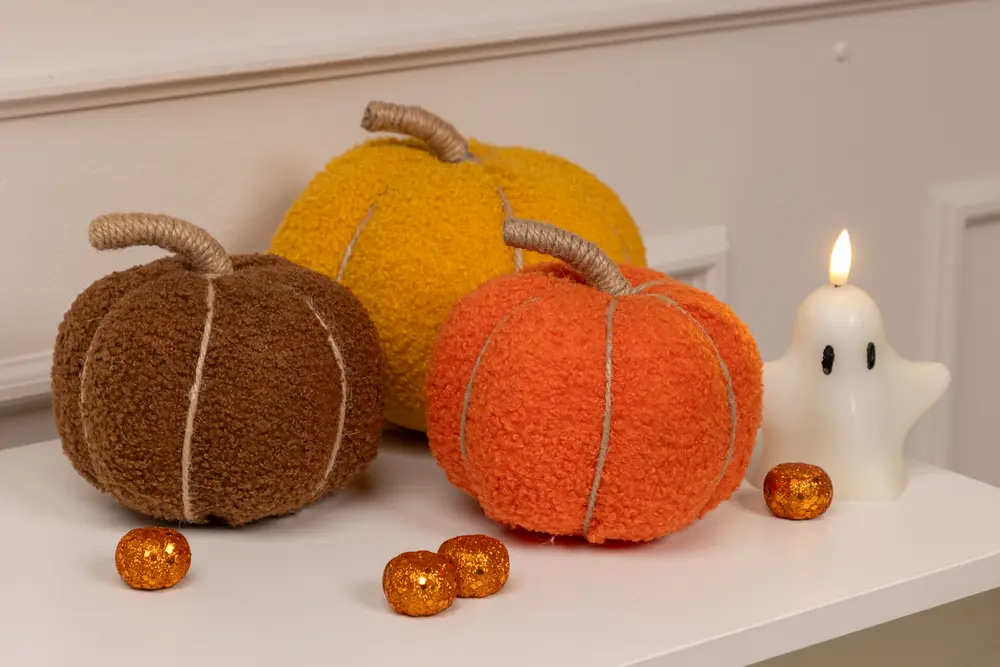 Three plush, textured pumpkins in brown, yellow, and orange colors are displayed on a white surface. Each pumpkin features a soft, fuzzy fabric with visible stitched lines and a thick, wrapped stem made of rope-like material. The pumpkins are accompanied by a small white ghost-shaped candle with black eyes and a lit flame, as well as several small, glittery orange decorative balls.