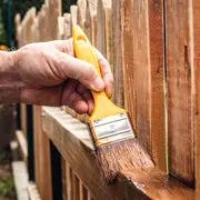 En hånd holder en pensel med gult håndtag og påfører træbeskyttelse eller maling på et hegn. Penslens børster og ergonomiske greb vises tydeligt.