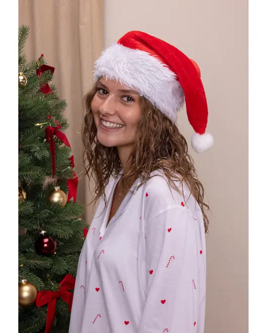 Hvid pyjamastop med røde hjerter og slikstokke, løs pasform, lange ærmer. Rød og hvid plyssanta-hue med kvast. Juletræ med pynt i baggrunden.
