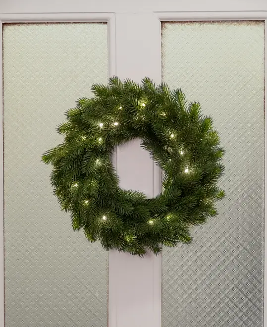 Kunstig grøn julekrans med naturtro grannåle, pyntet med små varme hvide LED-lys, velegnet til ophæng på døre eller vægge som juledekoration.
