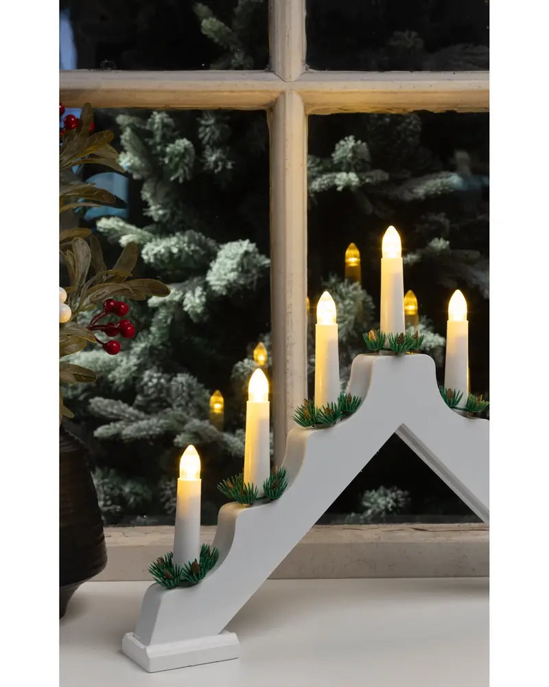 A white, wooden, triangular candle holder with seven electric candles arranged in a stepped pattern. Each candle has a warm yellow flame-shaped bulb and is decorated at the base with small artificial green pine sprigs. The candle holder sits on a windowsill, with a festive background including a Christmas tree and a plant with red berries.