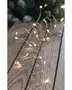 Thin, flexible silver wire string with small, warm white LED lights spaced evenly along its length. The wire is arranged in loose coils on a rustic wooden surface, showing its bendable nature. The lights emit a soft, cozy glow, ideal for decorative purposes.