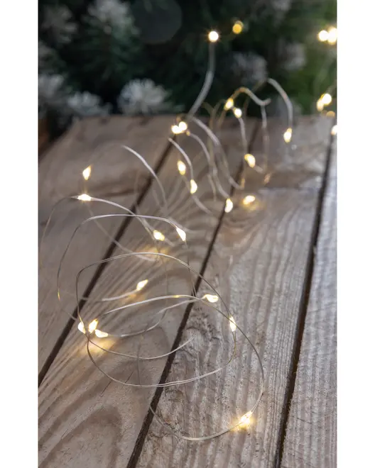 Thin, flexible silver wire string with small, warm white LED lights spaced evenly along its length. The wire is arranged in loose coils on a rustic wooden surface, showing its bendable nature. The lights emit a soft, cozy glow, ideal for decorative purposes.