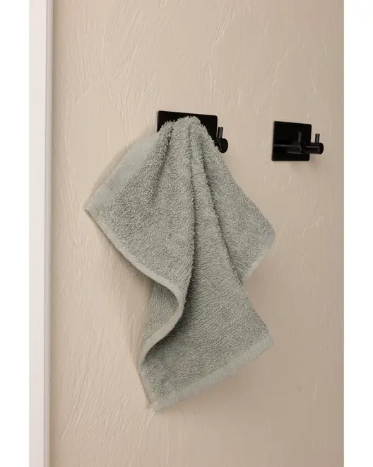 Lys grå håndklæde hænger på en moderne sort vægkrog, monteret på en beige væg med struktur. Håndklædet ser blødt og absorberende ud.