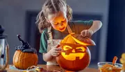 Barn med förkläde lyfter locket på en utskuren, lysande pumpa. Ytterligare en dekorerad pumpa och verktyg ligger på bordet, vilket tyder på Halloween-pumpakarvning.