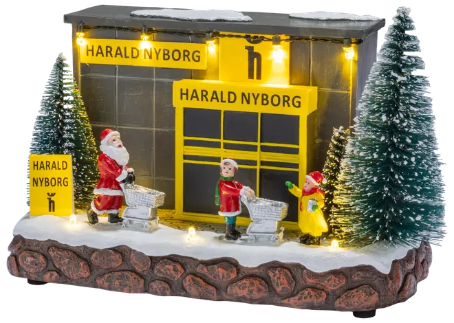 Festlig Harald Nyborg julelandsby med oplyst butik, nisse og børn-figurer, sneklædt base og snedækkede grantræer.