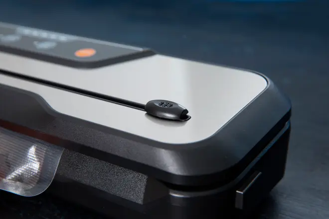 Nærbillede af en vakuumpakker med sort og sølv design, synlige kontrolknapper og forseglingsstrimmel. En delvist forseglet plastpose ses i kanten.