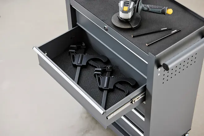 Grå metal værktøjskab med åben skuffe indeholdende to tvinger. Toppen har en slibemaskine og to skruetrækkere. Skuffen har en tekstureret måtte til beskyttelse af værktøj.