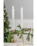Två vita kronljus i en guldfärgad ljusstake, dekorerad med gröna grankvistar och ett flätat hjärta, perfekt som julig bordsdekoration.