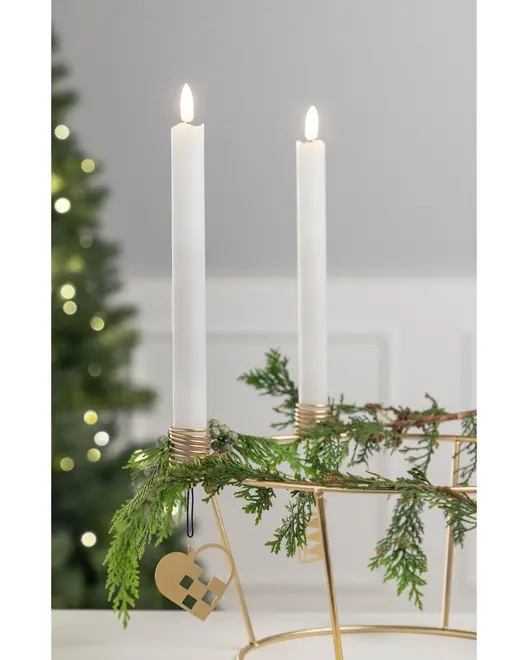 Två vita kronljus i en guldfärgad ljusstake, dekorerad med gröna grankvistar och ett flätat hjärta, perfekt som julig bordsdekoration.