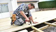 En person bruger en batteridrevet boremaskine til at montere terrassebrædder udendørs, iført værktøjsbælte med forskellige værktøjer, knælende på den under opførelse terrasse.