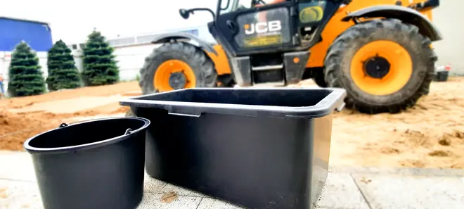 Sort plastikkasse og rund spand, begge med glatte overflader og solide kanter, velegnet til bygge- eller havearbejde. Vist udendørs på en arbejdsplads med maskine i baggrunden.