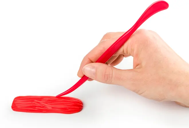 Rødt plastmodelleringsværktøj med buet håndtag, bruges til at forme ler eller modellervoks. Vist i hånd, presser tekstureret rød ler på hvid baggrund.