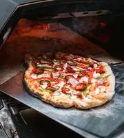 Træfyret pizza med sprød skorpe, ost, pepperoni, grøn og rød peber, tages ud af stenovn med en metal pizzaspade.