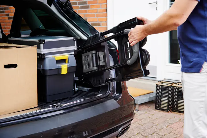 En person lægger en sammenklappelig sækkevogn i bilens bagagerum sammen med pap- og plastikkasser. Sækkevognen er sort med metalstel og kompakt design.