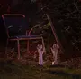 Halloween-trädgårdsdekoration med två realistiska plasthänder som sticker upp ur marken, bredvid en metallstol och en spade i en dunkelt belyst trädgård.