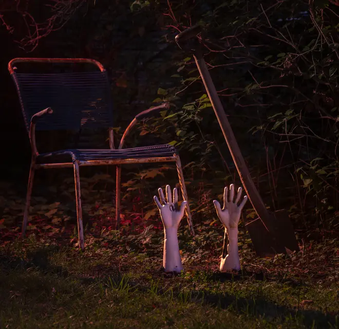 Halloween-trädgårdsdekoration med två realistiska plasthänder som sticker upp ur marken, bredvid en metallstol och en spade i en dunkelt belyst trädgård.