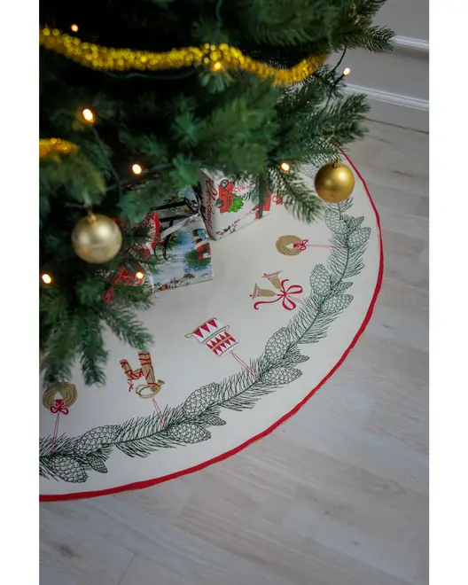 Rund julgransmatta med broderade grankvistar och festliga motiv, röd kant, placerad under granen med presenter. Krämfärgad bakgrund, dekorativ design för skydd av granens bas.