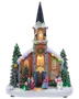 Oplyst julekirke med sneklædt spir, lys i vinduerne, grantræer, figurer, snemandsdekoration og farverige festlige lyskæder.