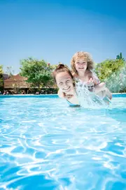 Utomhuspool med klart blått vatten, omgiven av grönska. Två barn leker och skvätter vatten i poolen under en solig himmel. Familjevänlig poolmiljö.