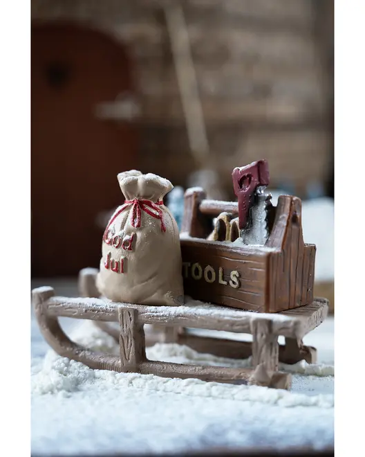 Julefigur med en træslæde, værktøjskasse med 'TOOLS' og en sæk med 'God Jul'-tekst, drysset med kunstig sne, perfekt til juledekoration.