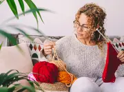 Kvinde strikker med rødt garn, holder strikkepinde og en kurv med rødt, orange, gult og pink garn, siddende på en sofa med mønstrede puder.