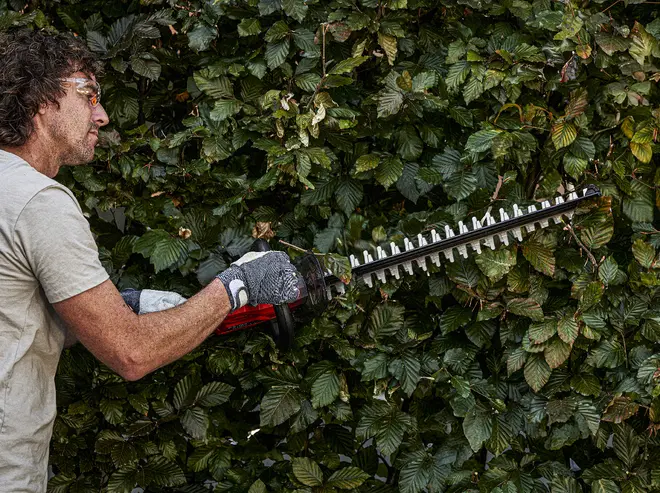 Elektrisk häcksax med långt dubbelsidigt blad, ergonomiskt handtag och skydd. Passar för klippning av häckar och buskar. Visas i bruk med skyddshandskar.