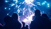 En gruppe mennesker ser et farverigt blåt og hvidt fyrværkeri på nattehimlen, med silhouetter synlige i forgrunden, hvilket skaber en festlig stemning.